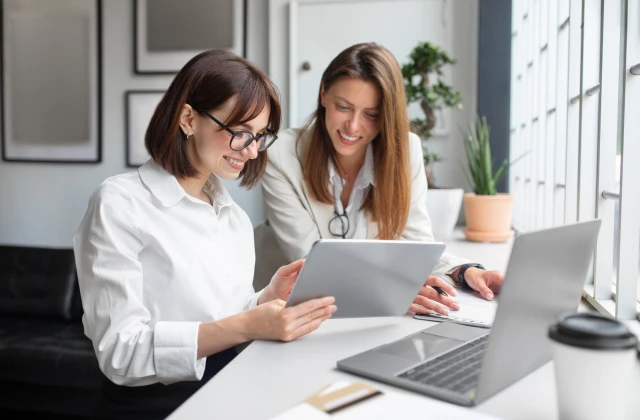 Two professional women in white shirts are working together in a modern office space. One woman, wearing glasses, is holding a tablet and smiling, while the other is looking at a laptop with a focused expression. The setting includes a bright workspace with large windows, framed pictures on the wall, and a coffee cup on the desk.