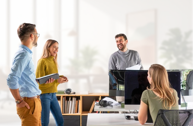 A small team collaborates around a desk in a modern office, discussing work displayed on a computer monitor.